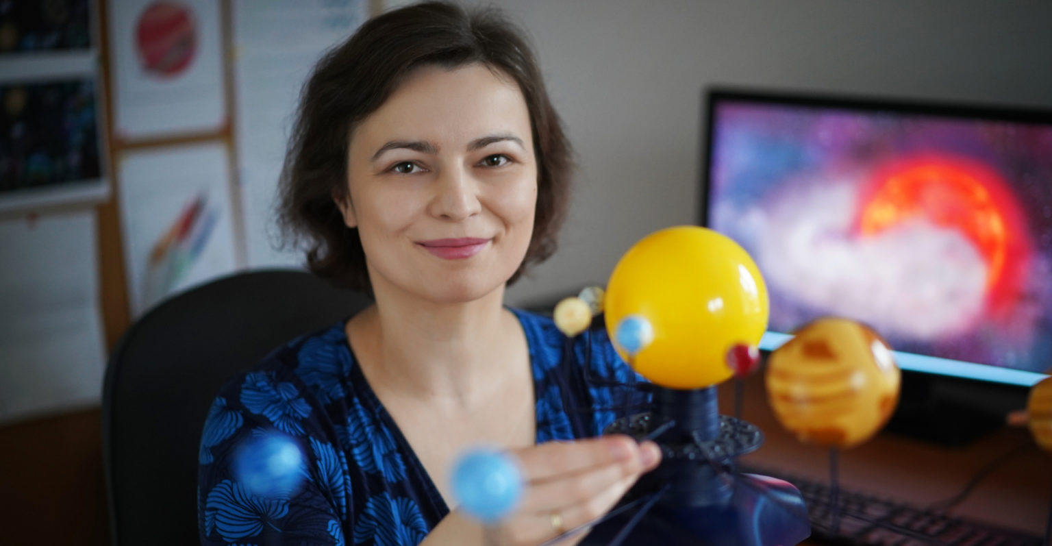 Kobieta w granatowej sukience trzyma w ręku żółte i niebieskie kule symbolizujące planety