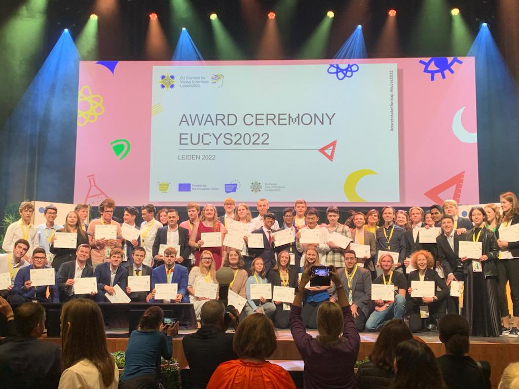 Na scenie stoi duża grupa finalistów europejskiego konkursu dla młodych naukowców, w rękach prezentują dyplomy