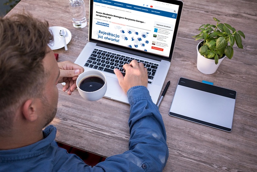 Mężczyzna patrzy na ekran laptopa i trzyma w ręku filiżankę z kawą. Na ekranie otwarta strona internetowa KPK