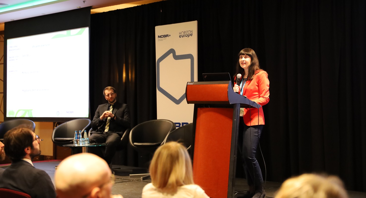 Na scenie za mównicą przemawia kobieta w czerwonej marynarce, w tle rolap \ konturem Polski i mężczyzna siedzący na fotelu. Na pierwszym planie głowy słuchaczy