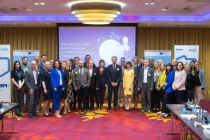 Na czerwonym dywanie stoi grupa kolorowo ubranych ludzi. W tle duży ekran z nazwą konferencji