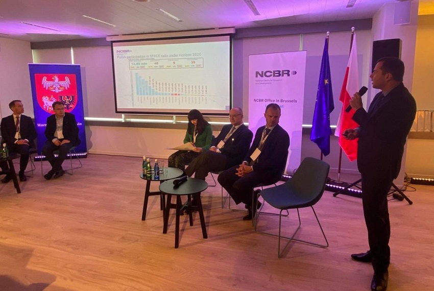 Na pierwszym planie stoi mężczyzna w garniturze mówiący do mikrofonu. Za nim na fotelikach siedzi pięcioro prelegentów. W tle ekran z prezentacją.