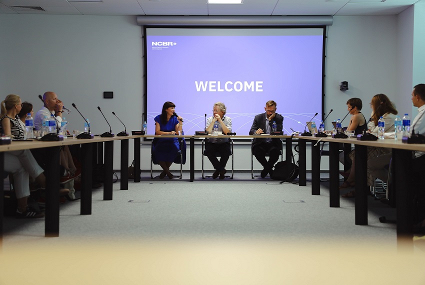 Przy stole ustawionym w podkowę siedzą uczestnicy spotkania. U szczytu stołu od lewej siedzi ciemnowłosa kobieta, jasnowłoasy mężczyzna w okularach i drugi mężczyzna w okularach i garniturze.