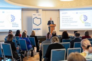 Sala konferencyjna wypełniona uczestnikami konferencji, z mównicy przemawia mężczyzna w garniturze