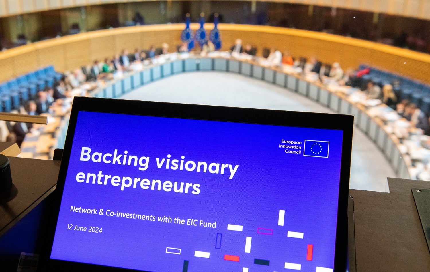 Na pierwszym planie ekran laptopa z napisem Backing visionary enterpreneurs, a w tle ludzie usadzeni w półkolu przy stole. Za nimi flagi Unii Europejskiej: niebieskie z żółtymi gwiazdkami