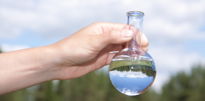 Czysta woda w okrągłym, szklanym naczyniu jest trzymana przez rękę. W tle zielone drzewa i niebo