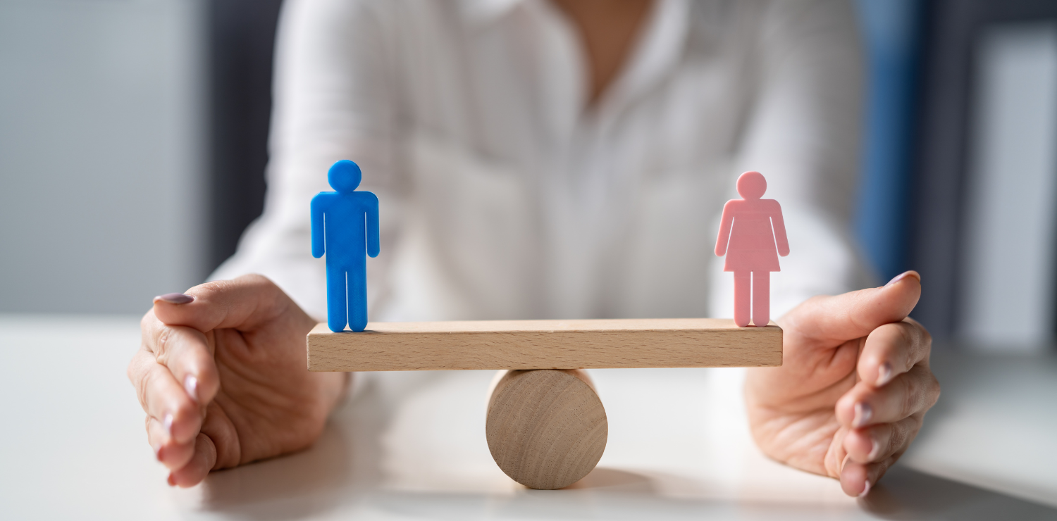 Na pierwszym planie drewniana huśtawka, z lewej stoi niebieska figurka mężczyzny, z prawej różowa figurka kobiety. W tle korpus osoby w białej koszuli.