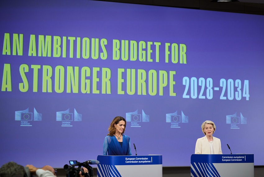 Na zdjęciu są dwie kobiety: blondynka w jasnej garsonce po prawej i szatynka w niebieskiej sukience po lewej. W tle wielki ekran z napisem 