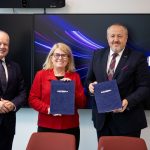 Na zdjęciu są trzy osoby: dwaj mężczyźni w ciemnych garniturach, w środku kobieta w czerwonym żakiecie. Kobieta i mężczyzna po prawo prezentują dokument w granatowej teczce z napisem NCBR.