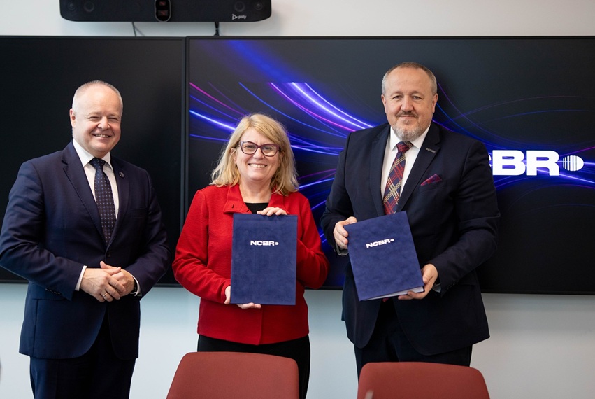 Na zdjęciu są trzy osoby: dwaj mężczyźni w ciemnych garniturach, w środku kobieta w czerwonym żakiecie. Kobieta i mężczyzna po prawo prezentują dokument w granatowej teczce z napisem NCBR.