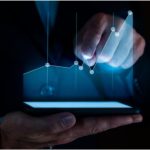 Z ciemności wyłaniają się męskie dłonie: jedna trzyma ekran, który emituje światło układające się w wykres, a druga wskazuje jeden z punktów tego wykresu.