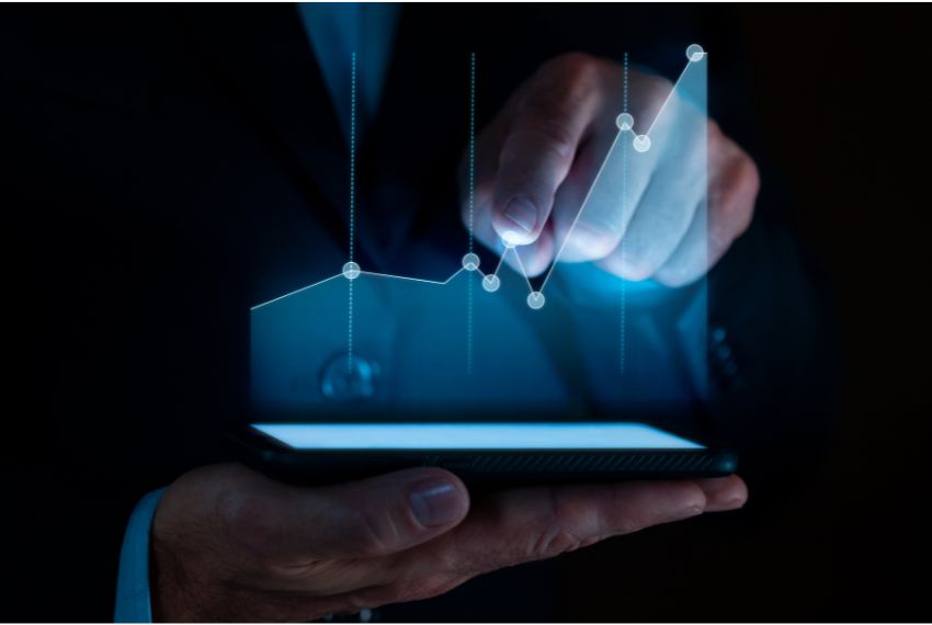 Z ciemności wyłaniają się męskie dłonie: jedna trzyma ekran, który emituje światło układające się w wykres, a druga wskazuje jeden z punktów tego wykresu.