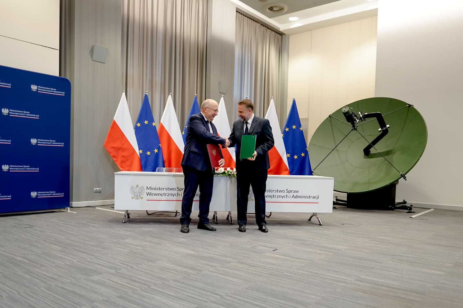 Dwaj mężczyźni w ciemnych garniturach ściskają sobie dłonie, dzierżąc w rękach podpisaną umową. W tle biało-czerwone flagi Polski i granatowe flagi Unii Europejskiej oraz duża, okrągła zielona czasza anteny satelitarnej
